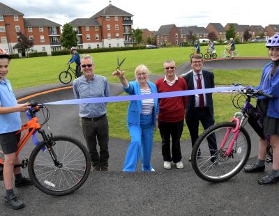 Image (l-r): Ward councillor, Paul Warburton; deputy council leader, Jean Flaherty; Great Sankey parish councillor, Steve Parish; and the council’s director for transport and environment, David Boyer, preparing to cut a ribbon at the new pump track in Great Sankey. They are surrounded by smiling primary school children on bicycles.