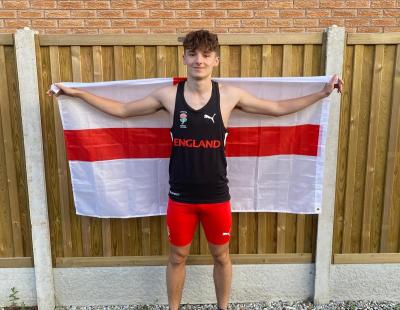 Image of Lewis Shaw, a member of LiveWire’s Talented Athletes programme. He is standing in front of a fence in full athletics kit. He has an England flag stretched out over his shoulders and behind his back.