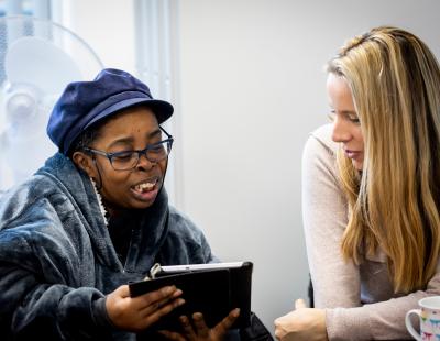 Image of two people talking, one person is holding up a tablet device to the other and both are looking at this. 